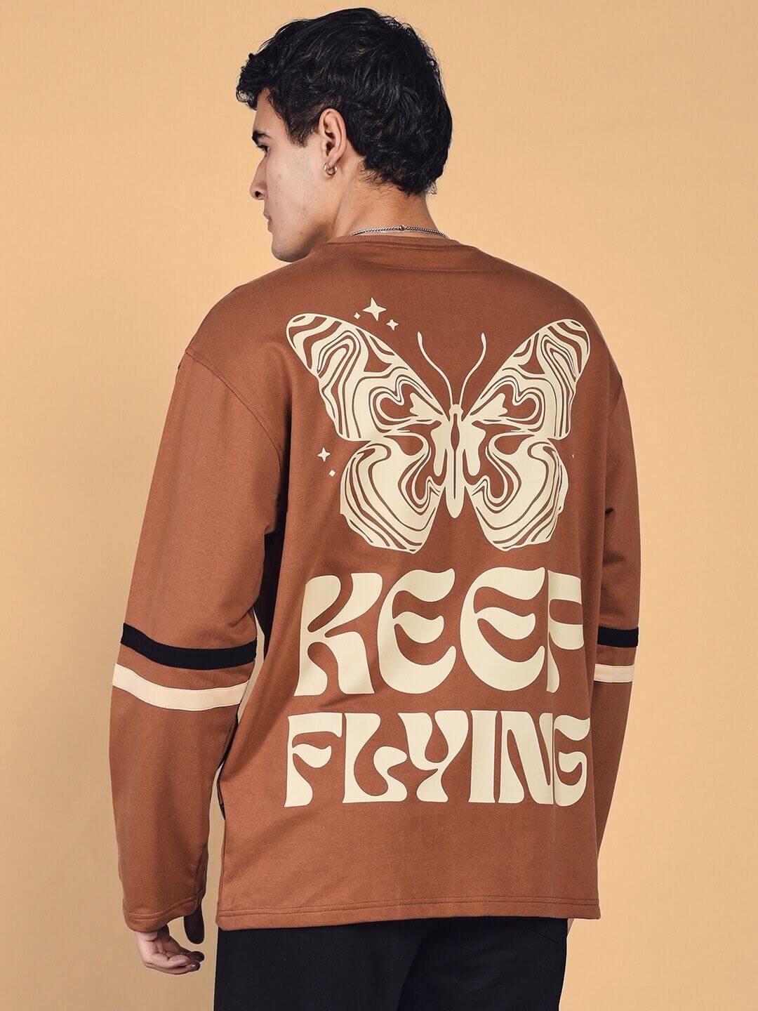 Man wearing brown oversized sweatshirt with 'Keep Flying' and butterfly design.