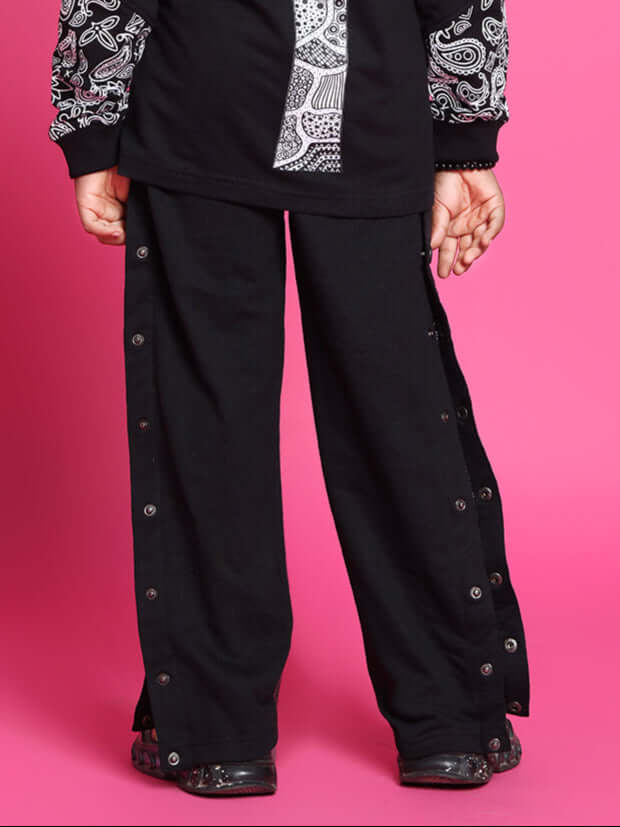 Child wearing black motif joggers with button details, standing against a pink background.