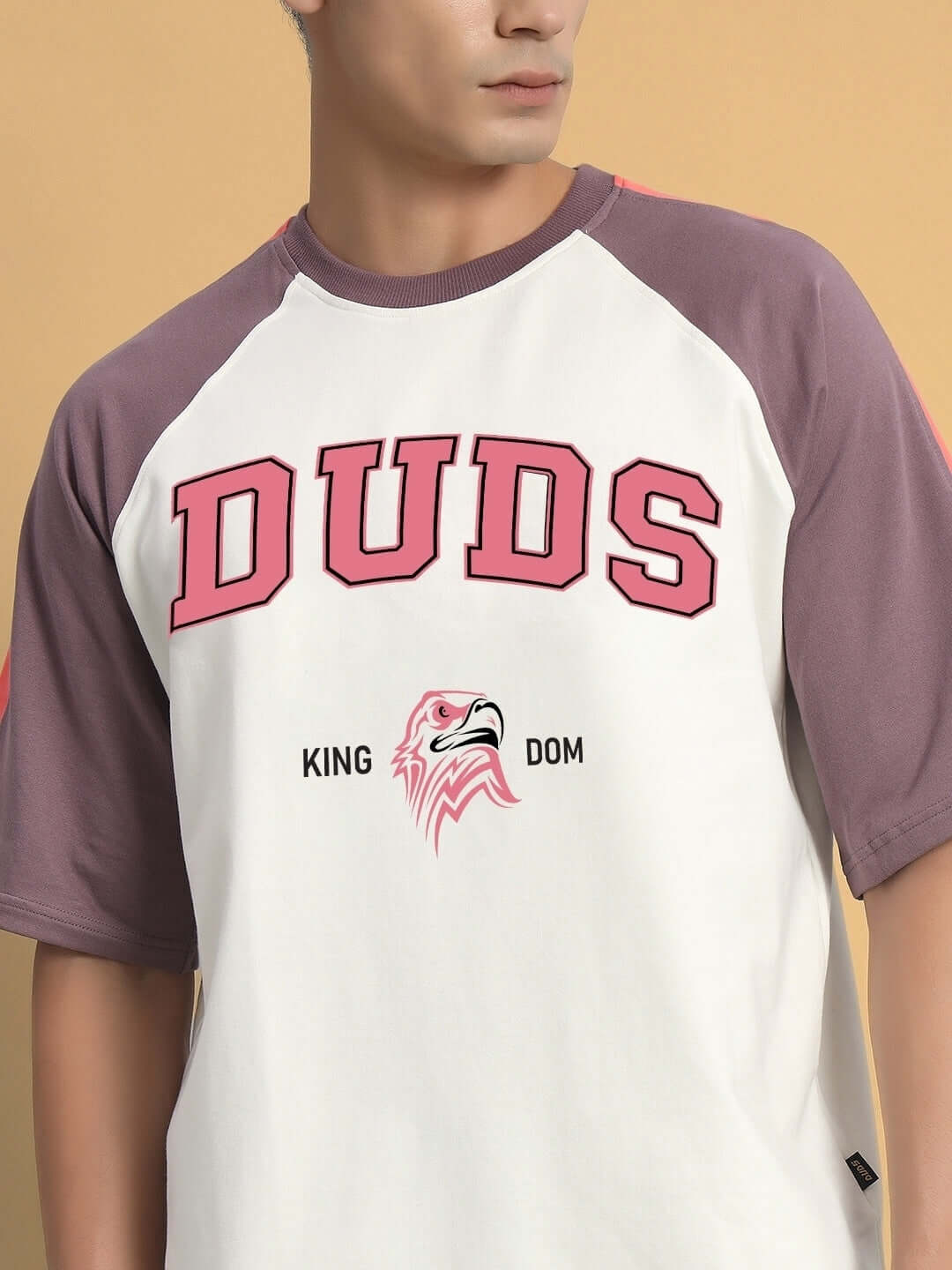 Model wearing KINGDOM Raglan Blocked Oversized T-Shirt in off white and violet, featuring bold 'DUDS' text and eagle graphic.