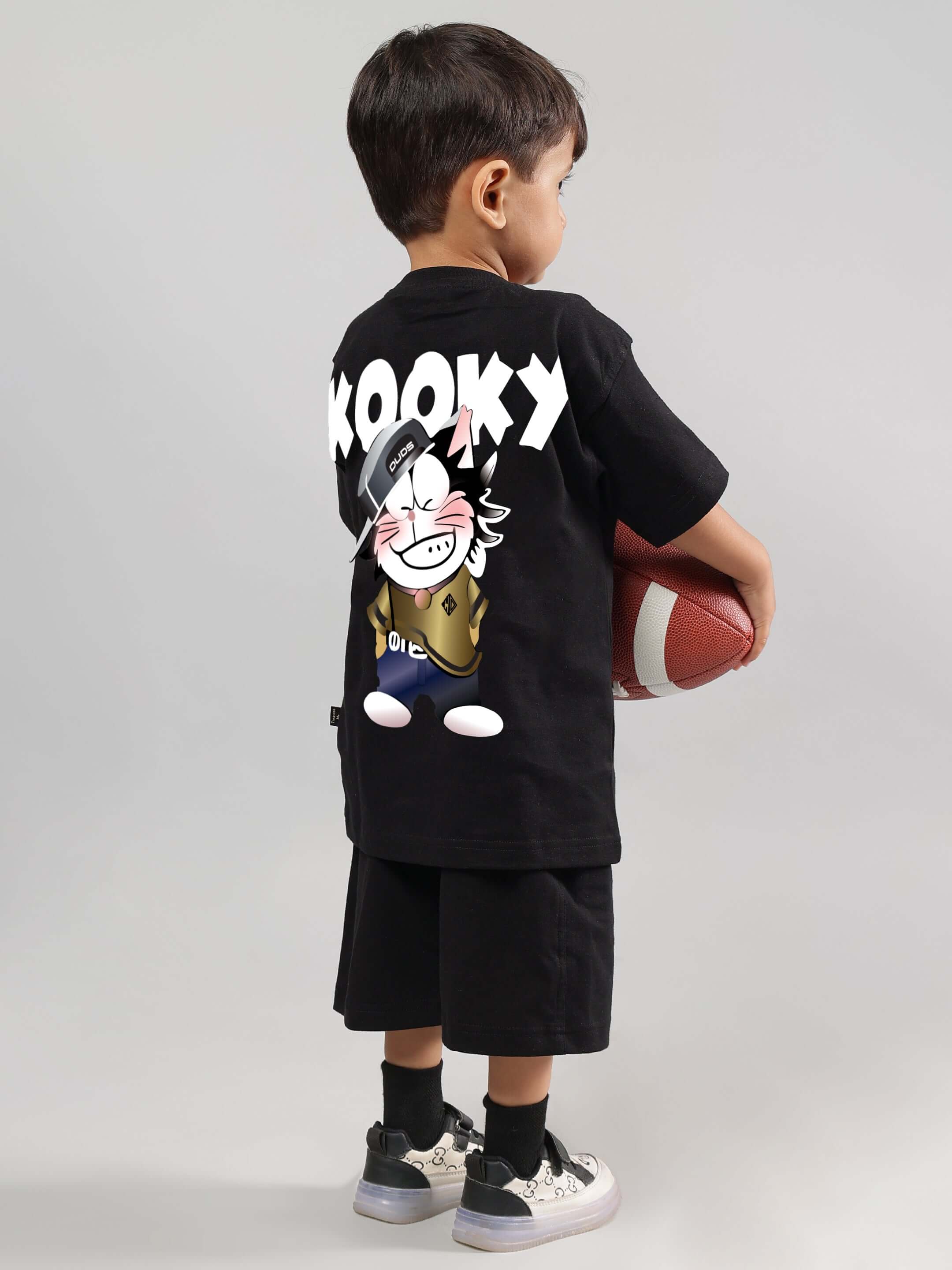 Child wearing KOOKY black co-ord set holding a football, showcasing cartoon design on back. Stylish attire for boys and girls, available in custom fit.