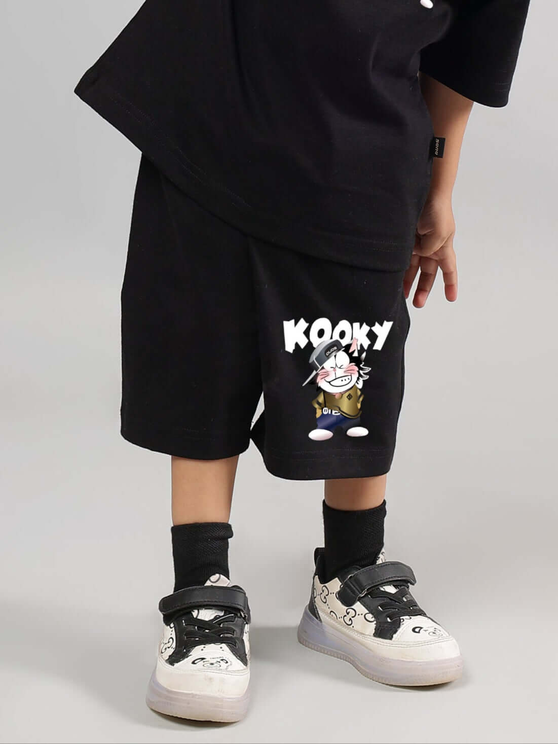 Child wearing black KOOKY shorts with graphic print, standing indoors in designer sneakers.