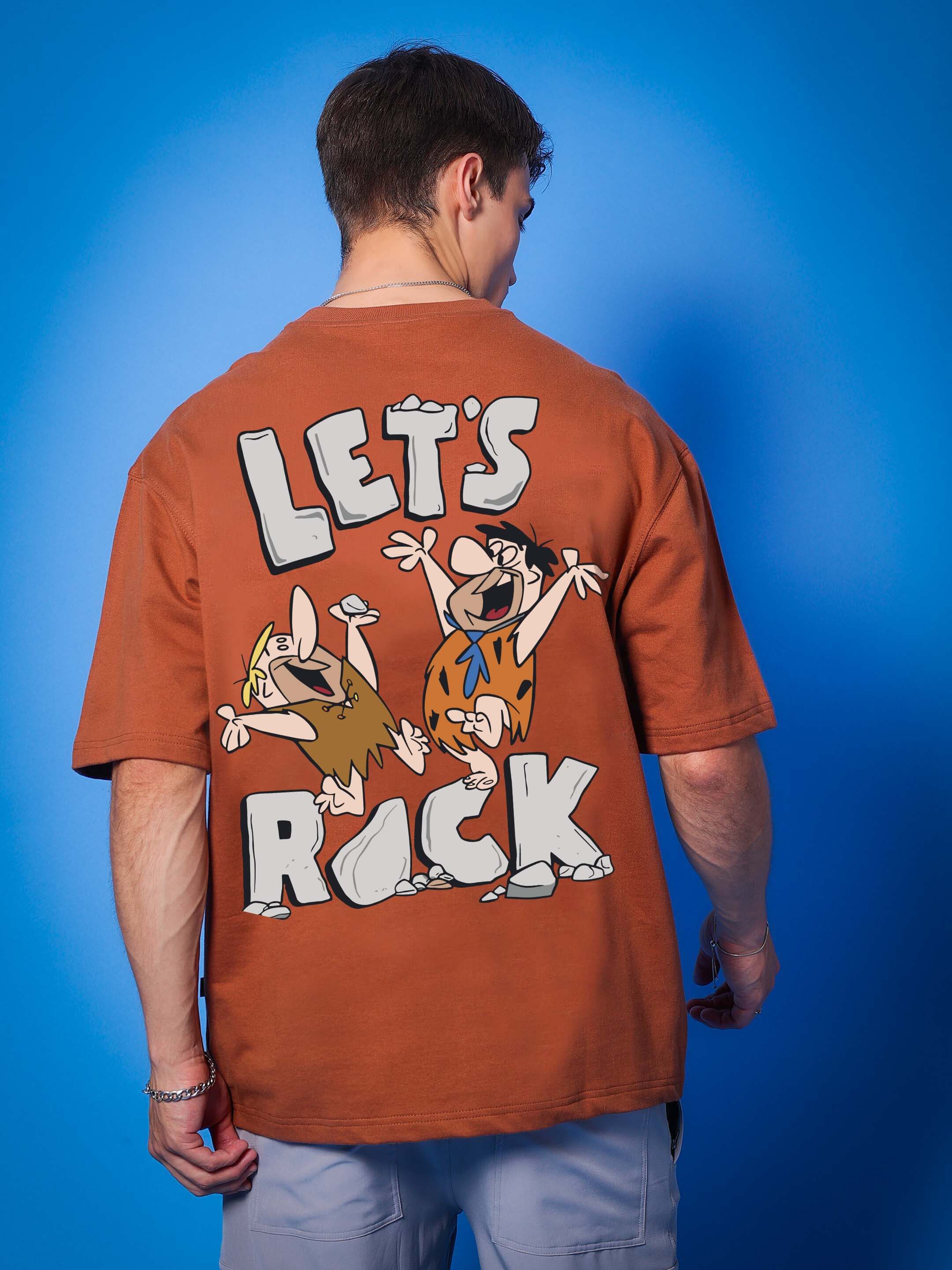 Model wearing brown "LET'S ROCK" oversized T-shirt with cartoon graphics, made of 97% cotton French terry, perfect for casual wear.