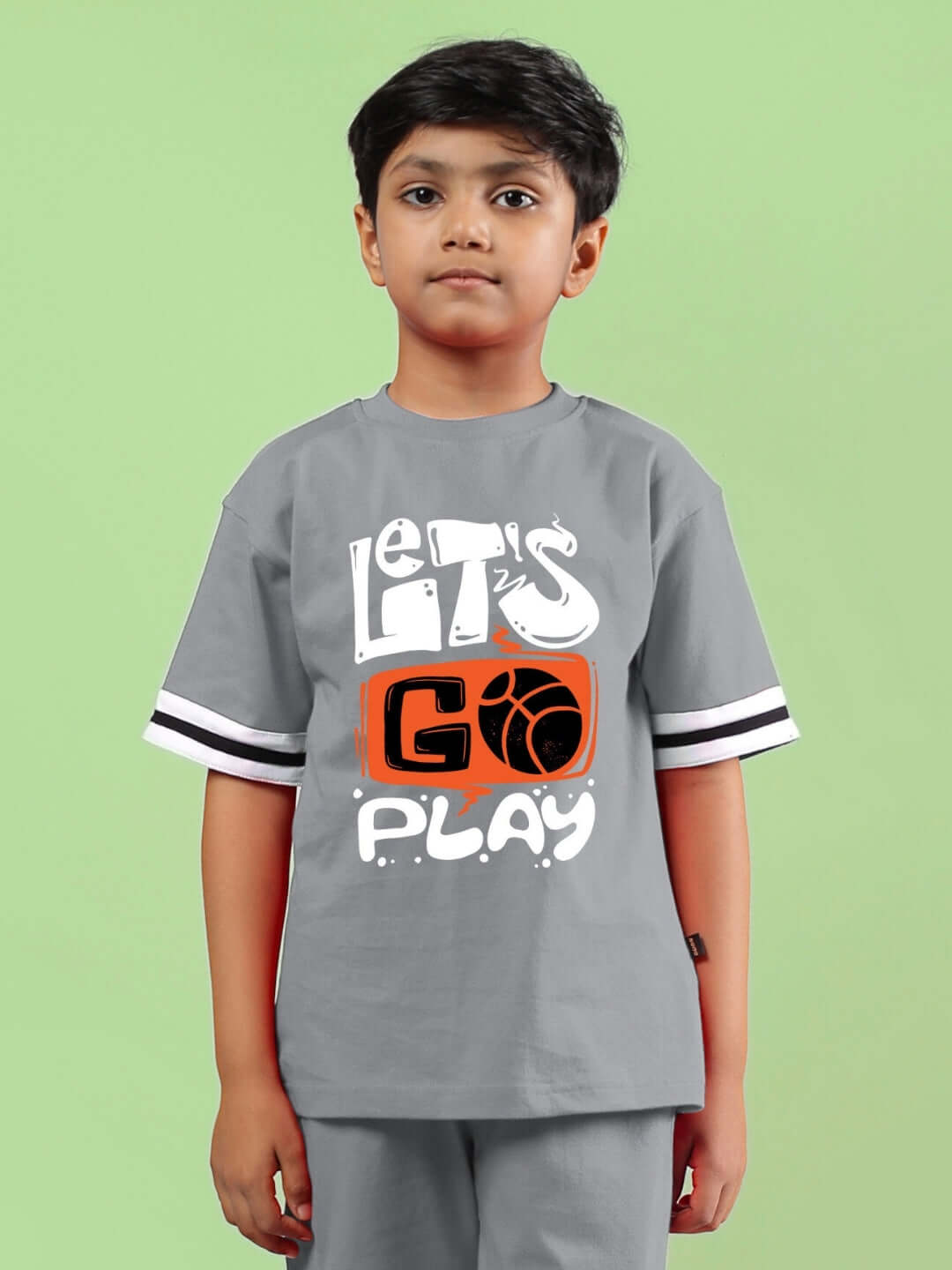 Boy wearing a grey 'Let's Go Play' printed t-shirt, designed for both boys and girls, in a casual setting against a green background.