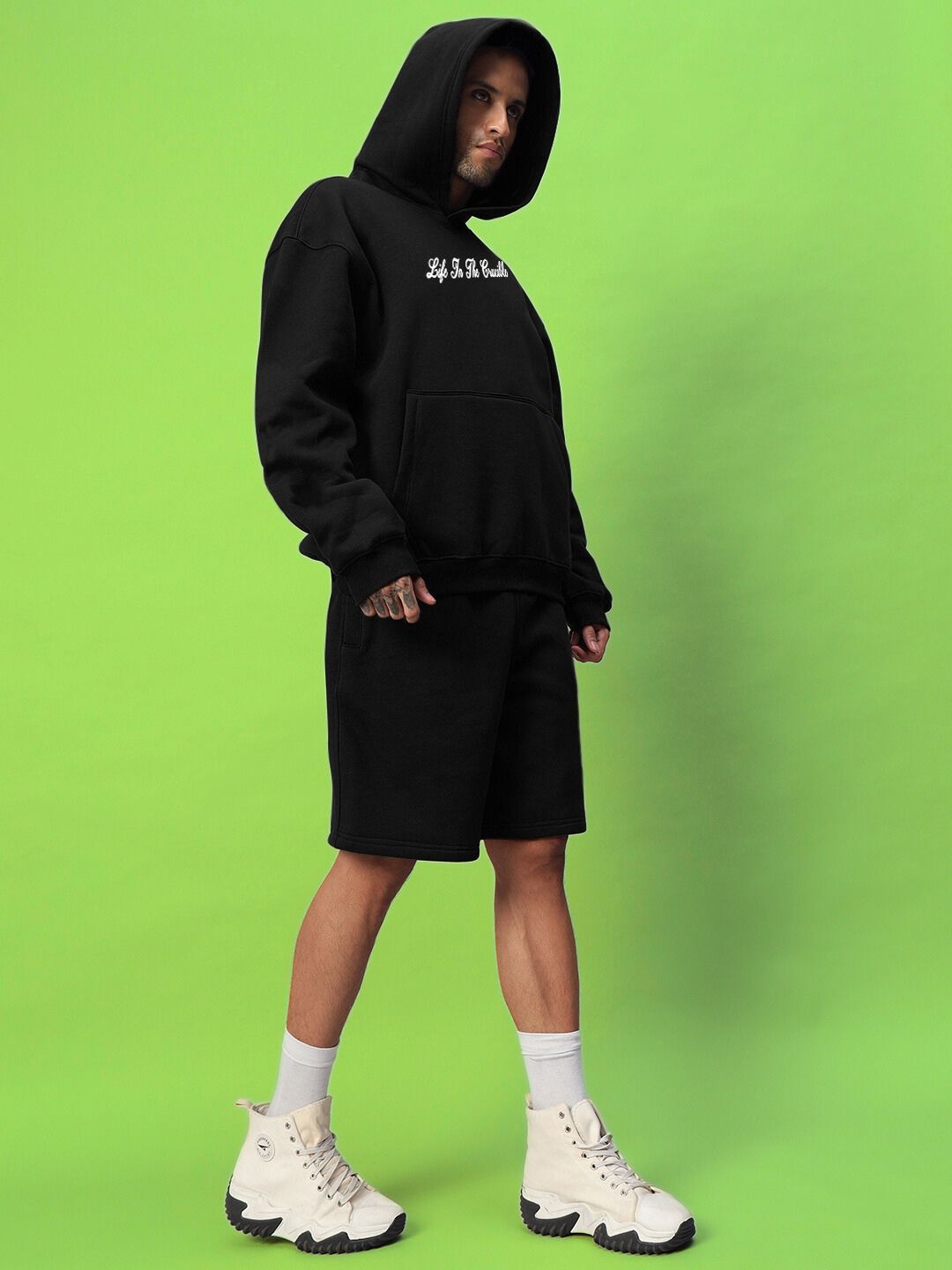 Model wearing LIFE IN THE CRUCIBLE PRINTED FLEECE CO-ORD BLACK on green background, showcasing streetwear style with graphic print.