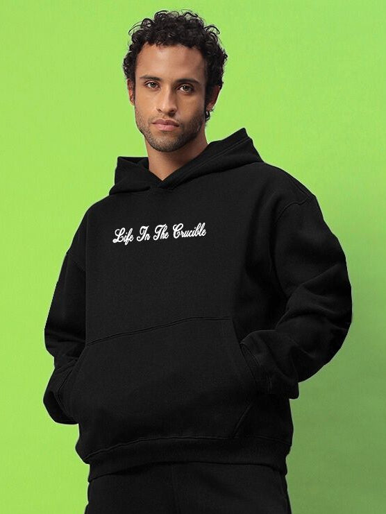 Model wearing LIFE IN THE CRUCIBLE PRINTED FLEECE HOODIE BLACK against green background, showcasing modern streetwear style.