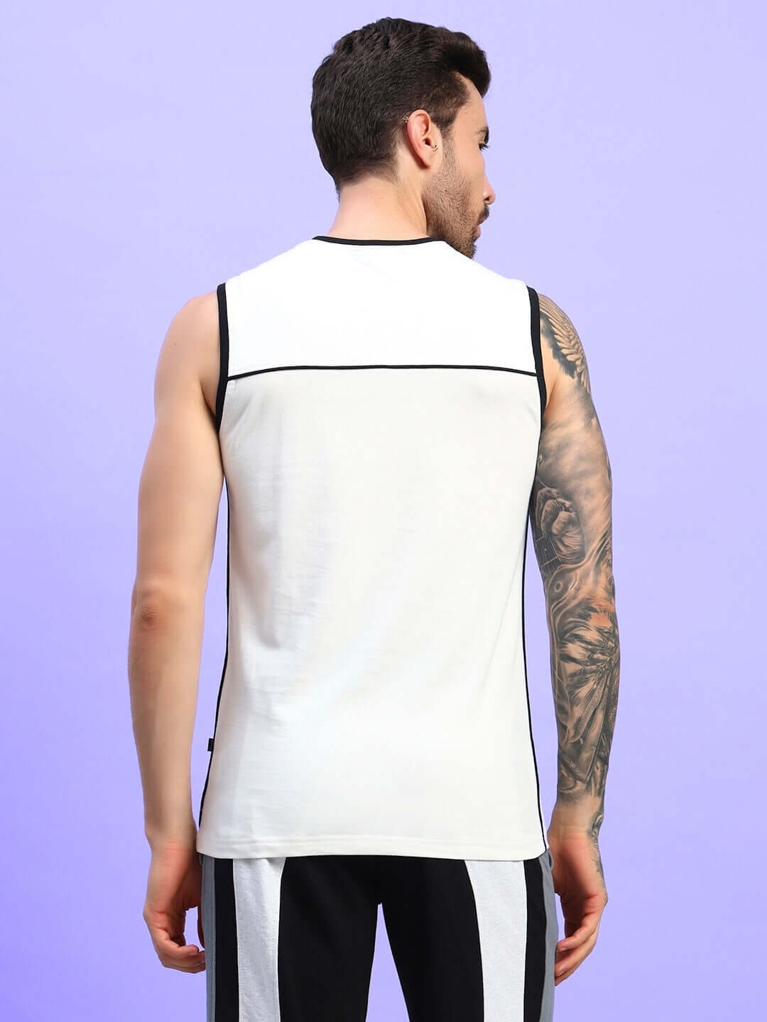 Man wearing a sleeveless white and black panel t-shirt with tattoo, facing backward against purple background.