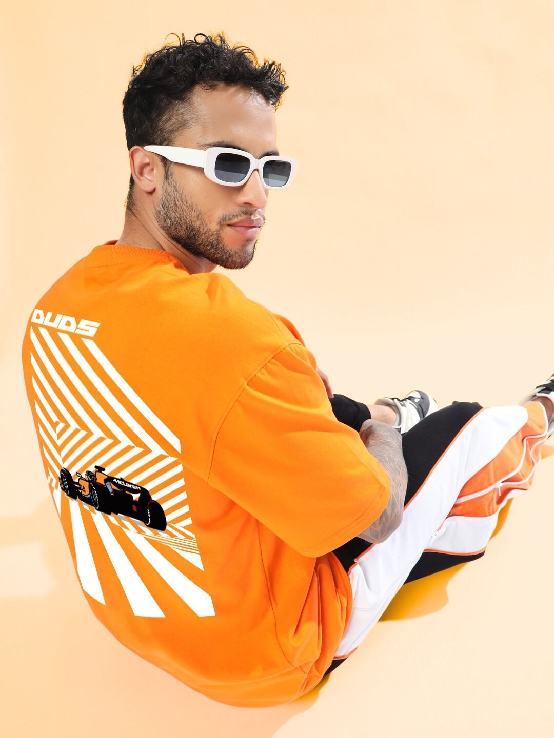 Model wearing MACLERAN PRINTED OVERSIZED T-SHIRT ORANGE, sitting on a peach background, showcasing back design, paired with sunglasses.