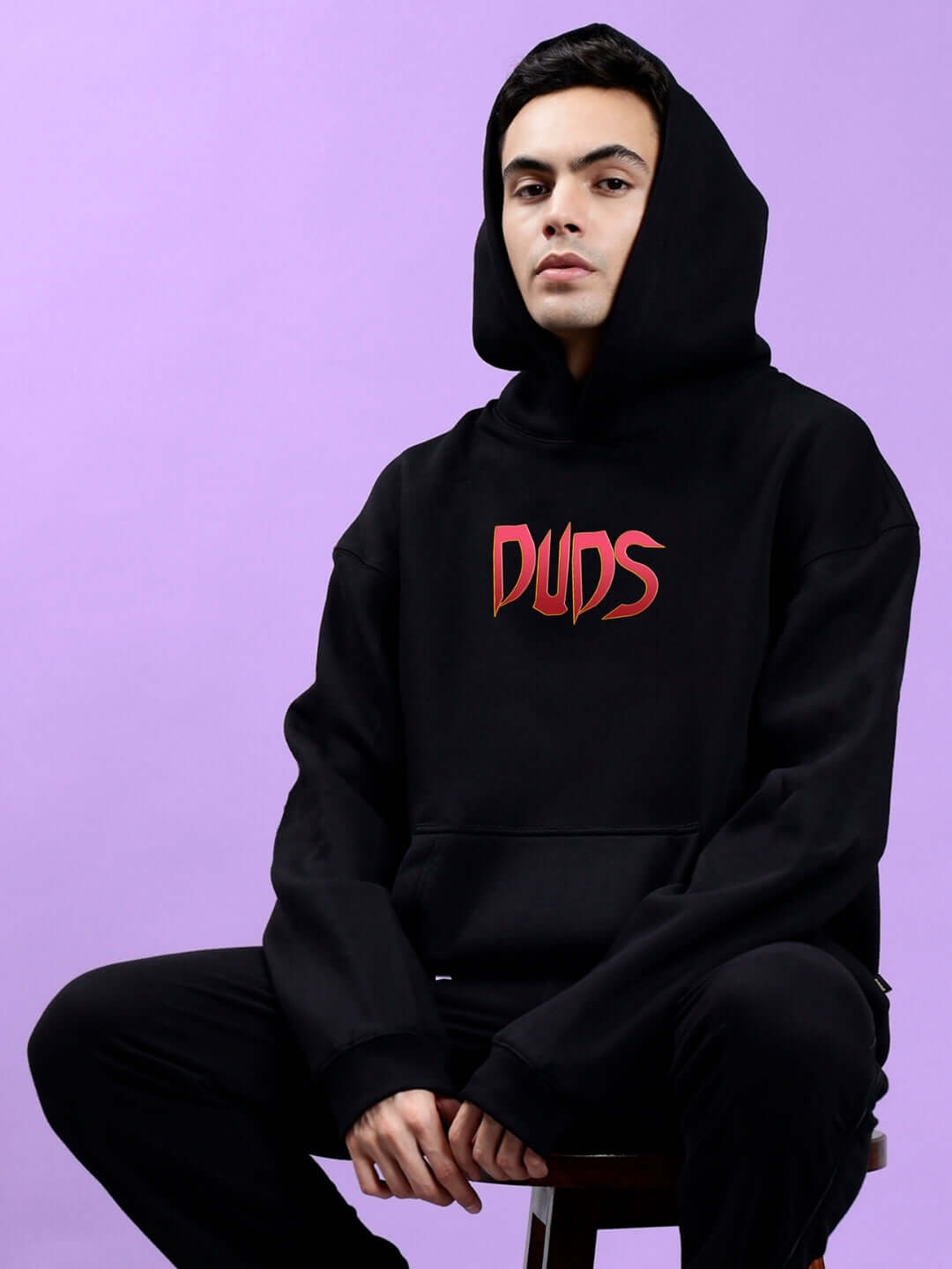 Person wearing MAFIA FLEECE OVERSIZE HOODIE BLACK with bold graphic, seated against purple background.