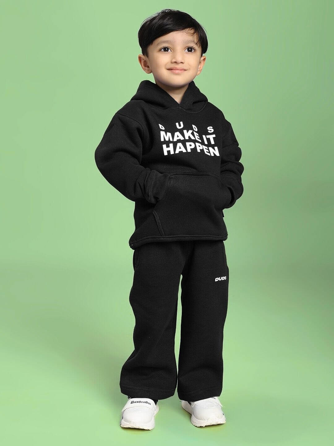 Young boy wearing MAKE IT HAPPEN FLEECE JOGGER FOR BOYS AND GIRLS BLACK with white sneakers standing on a green background.