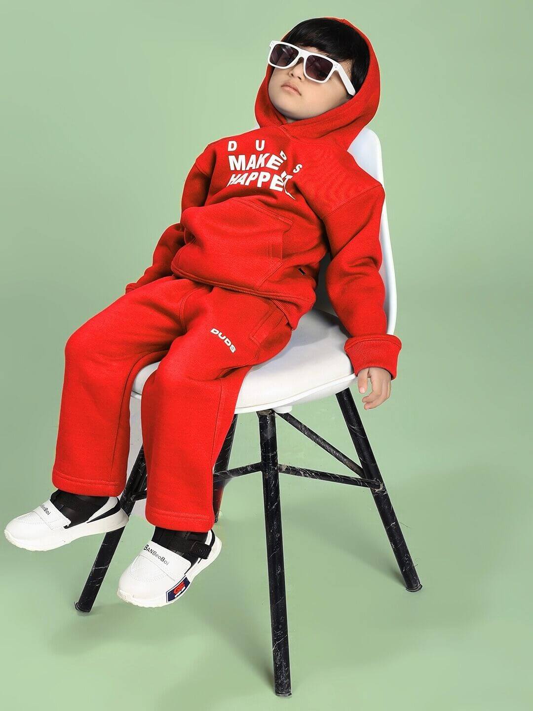 Child wearing MAKE IT HAPPEN FLEECE CO-ORD FOR BOYS AND GIRLS RED with sunglasses, sitting on a chair against a green background.