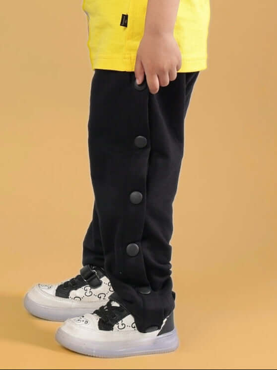 Child wearing Mambo Jogger in black-red with button detail, paired with graphic sneakers and a yellow shirt. Unisex, custom fit available.