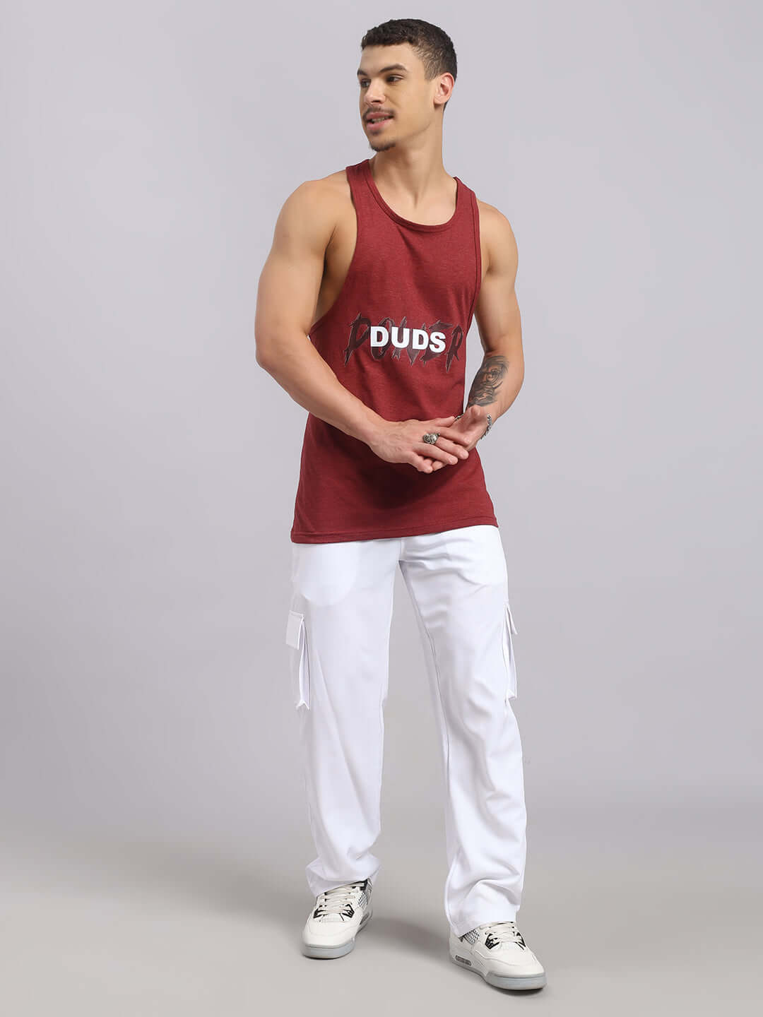 Man wearing a maroon printed gym tank top labeled DUDS paired with white cargo pants and sneakers, showcasing casual fitness wear.