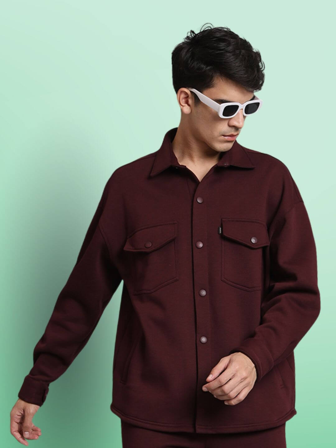 Man wearing maroon relaxed fit full sleeves fleece overshirt with white sunglasses, posing against a light green background.
