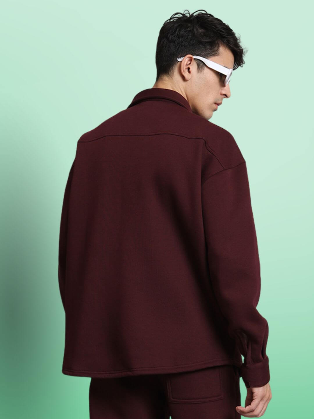 Model wearing maroon relaxed fit full sleeves fleece overshirt with sunglasses against a green background.