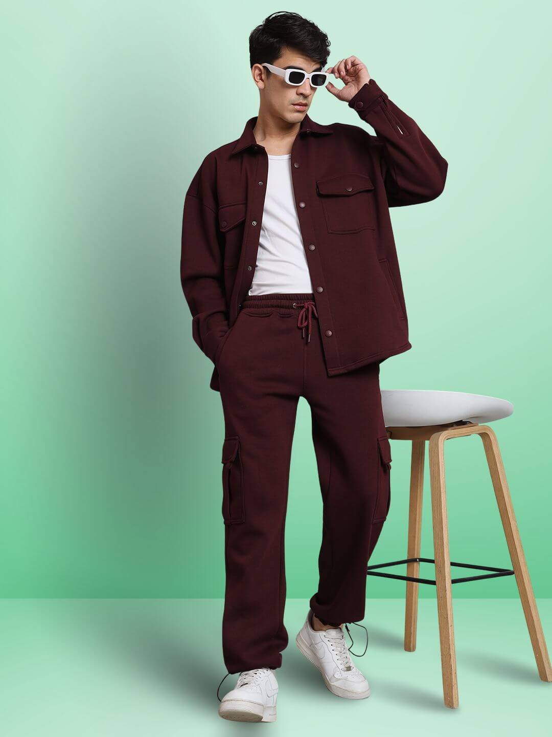 Man wearing maroon relaxed fit, full sleeves fleece overshirt with matching pants and white sneakers, posing in front of a chair.