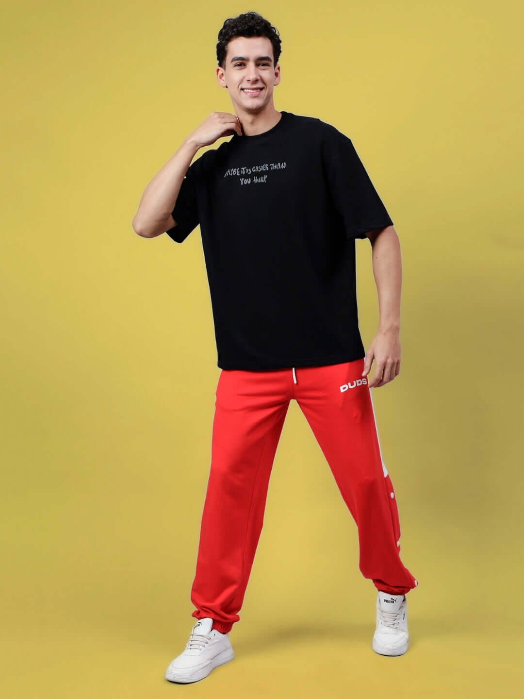 Man wearing oversized black t-shirt and red pants, styled with white sneakers, posing confidently against a yellow background.