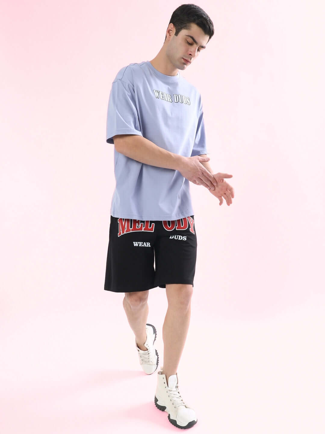 Man wearing black Melody Regular Fit Shorts with red and white logo and a light purple shirt against a pink background.