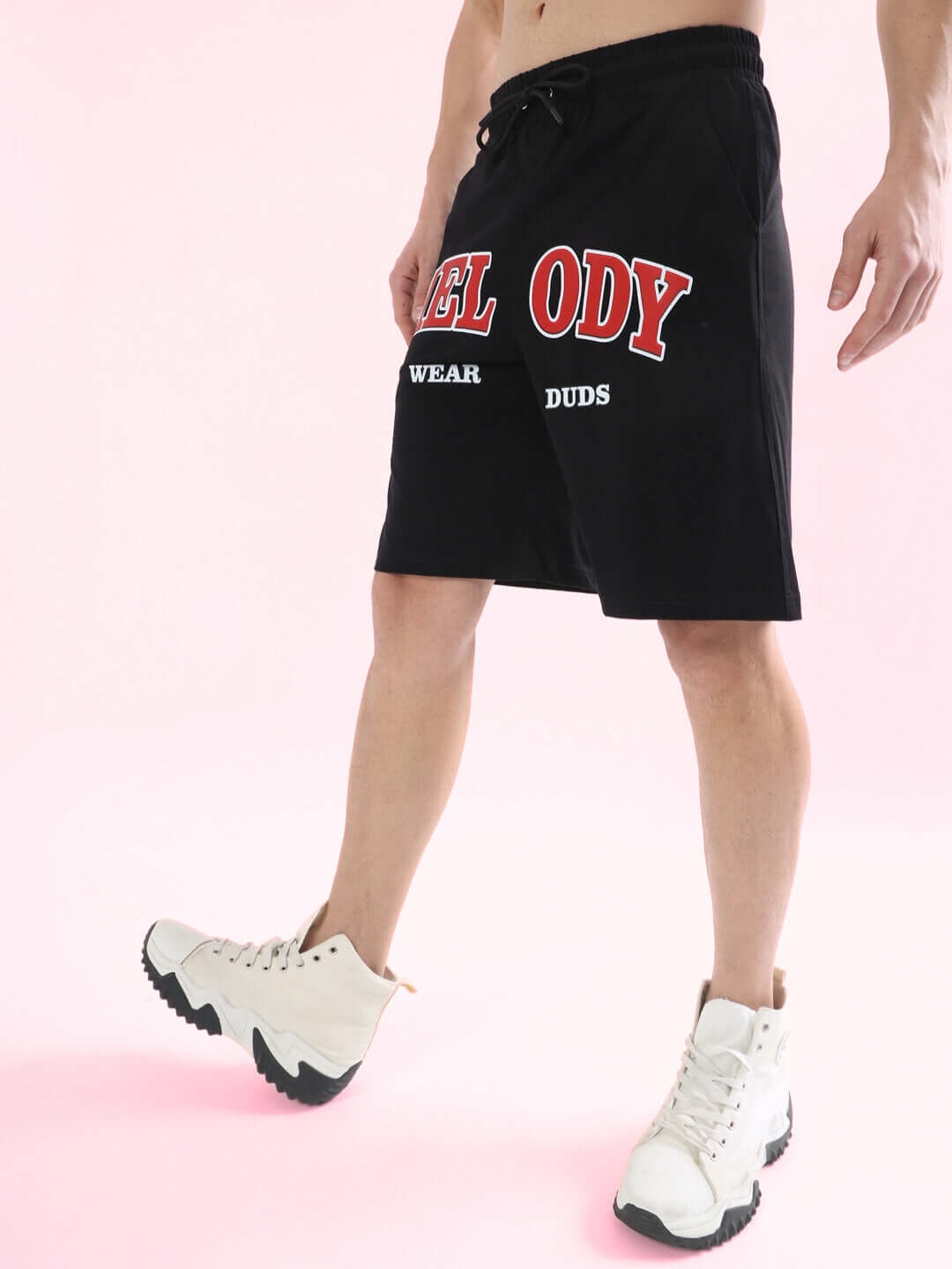 Person wearing black Melody regular fit shorts with red lettering and white sneakers, standing against a pink background.