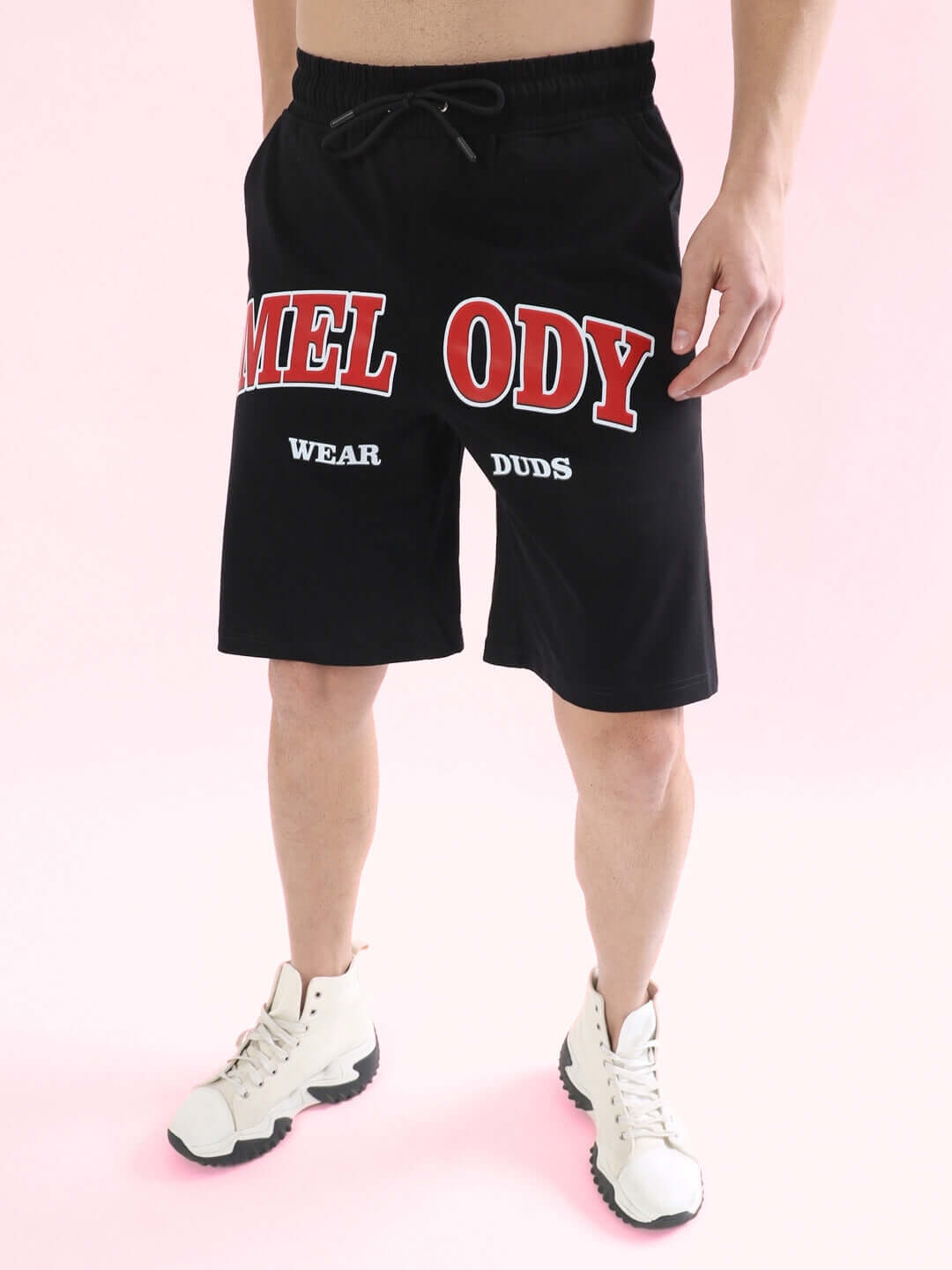 Person wearing black Melody regular fit shorts by Wear Duds, featuring bold red lettering, on a pink background.