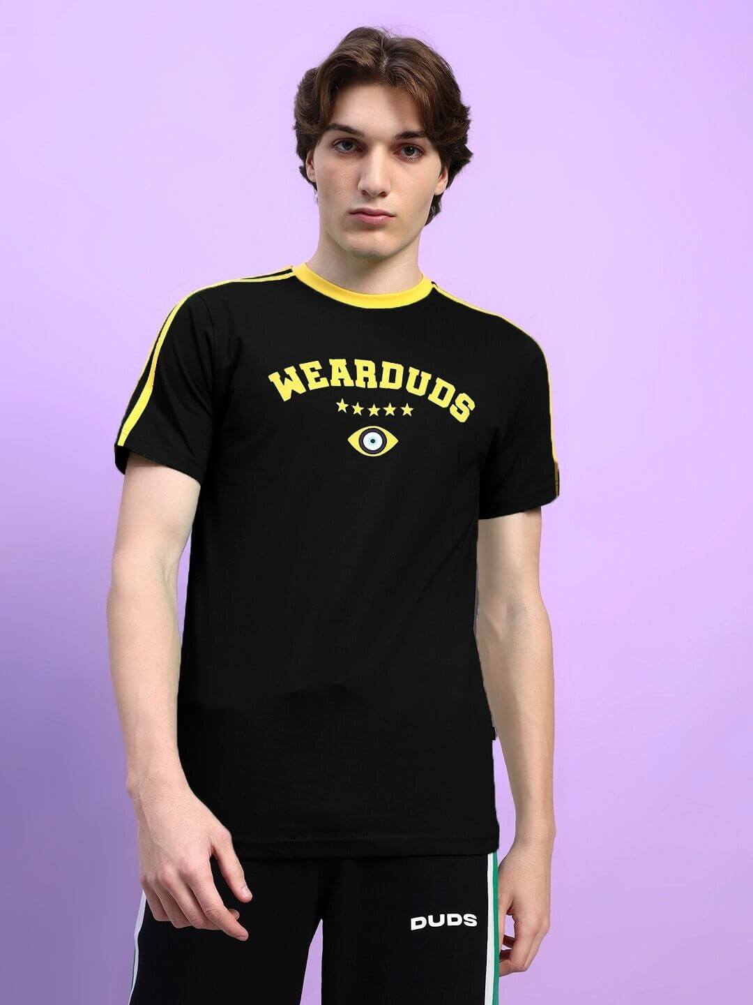 Model wearing a black Wearduds t-shirt with yellow accents featuring an eye design, standing against a purple background.