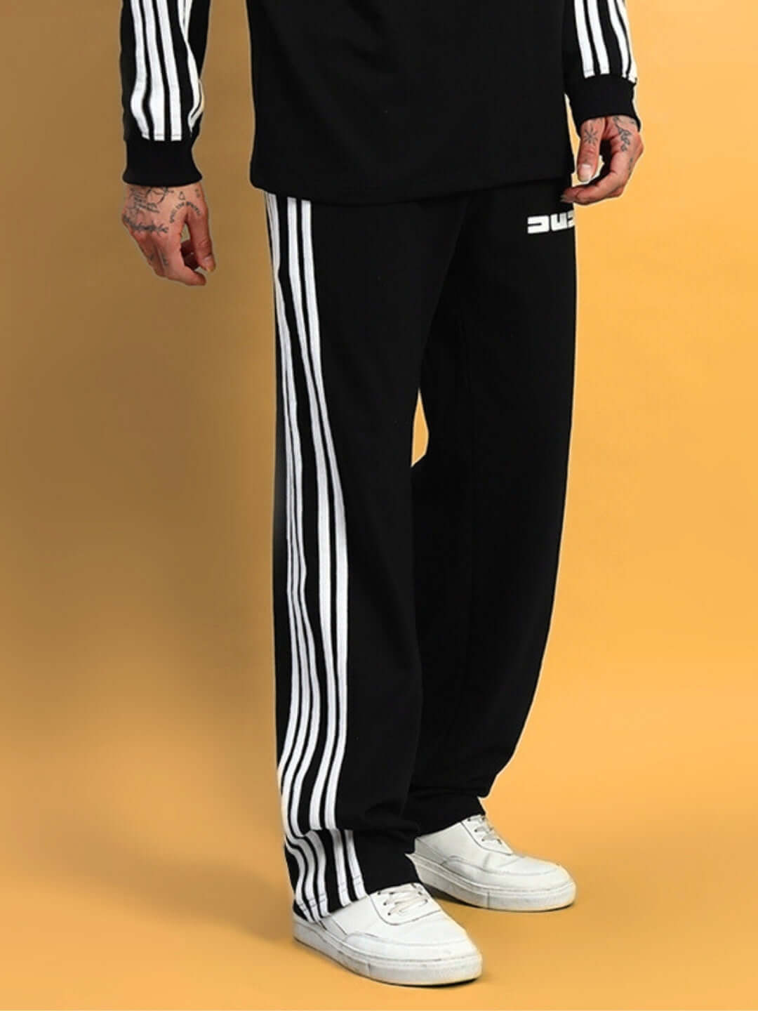 Black PARKA jogger pants with white stripes, worn by a person, showcasing casual sportswear style on a yellow background.