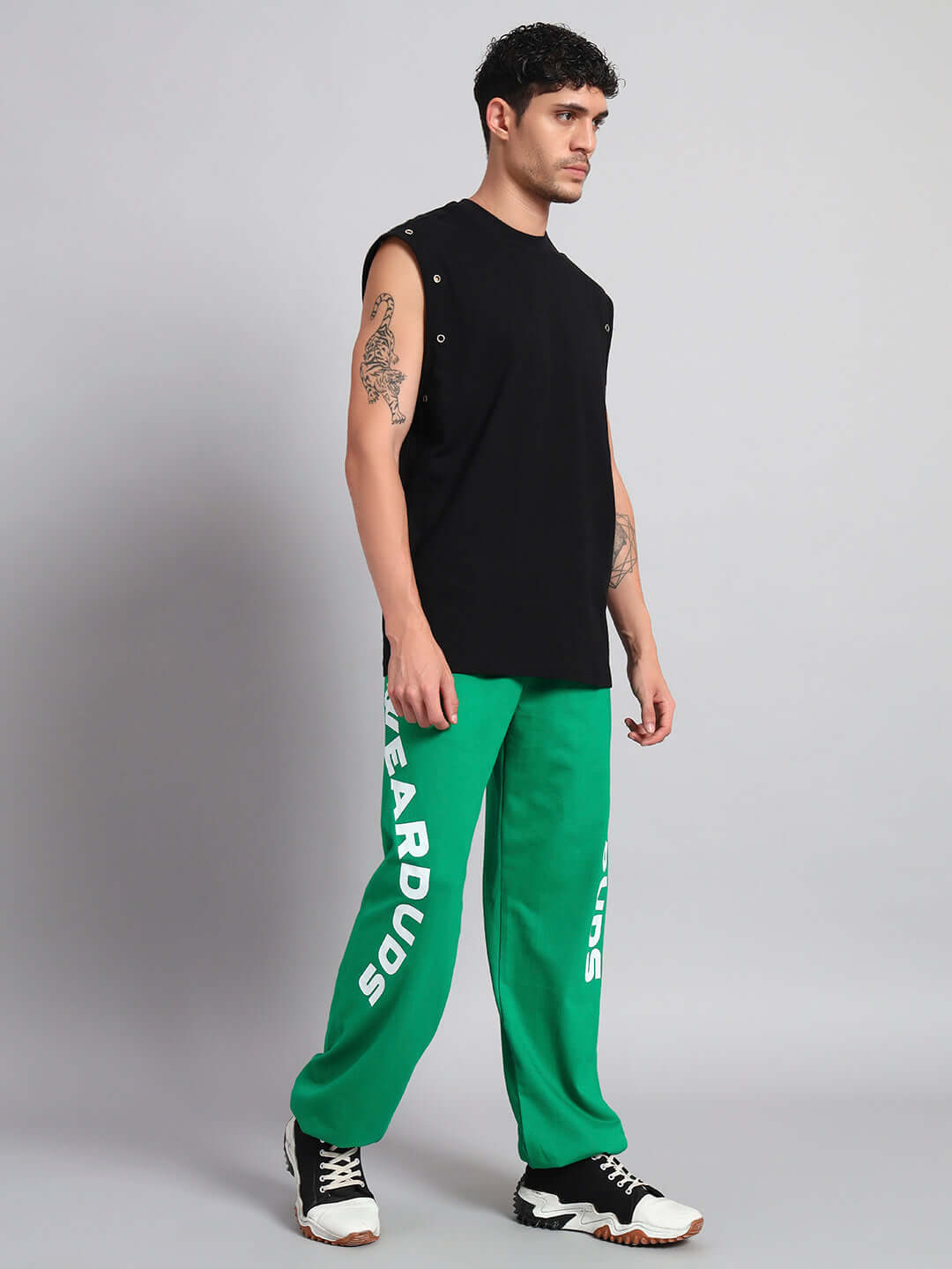Man posing in black muscle shirt and green sweatpants with sneakers on a neutral backdrop.