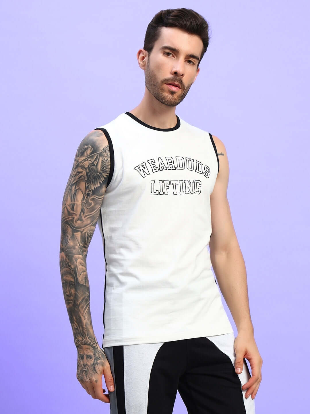 Man wearing a white sleeveless "Weardrdo Lifting" t-shirt with black trim, showcasing tattoos and black and white pants against a purple background.