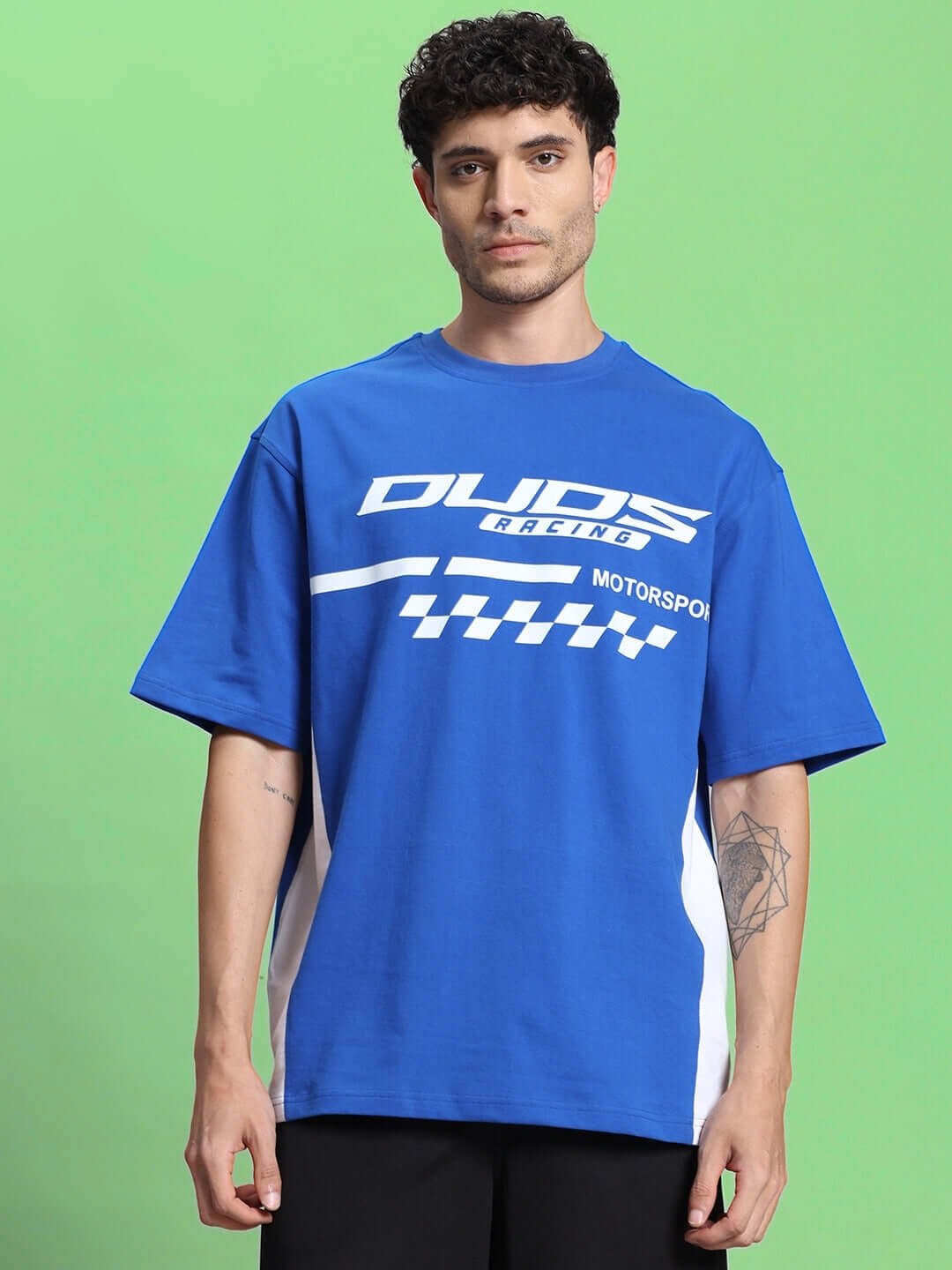 Model wearing INDY OVER-SIZED CLASSIC T-SHIRT ROYAL BLUE with checkered design and bold logo on green background.