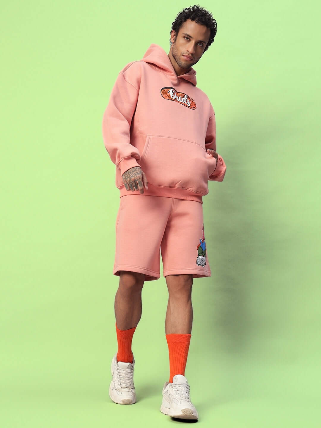 Man modeling DELIBERATE FLEECE CO-ORD PEACH, wearing a peach-colored hoodie and shorts with orange socks and white sneakers.