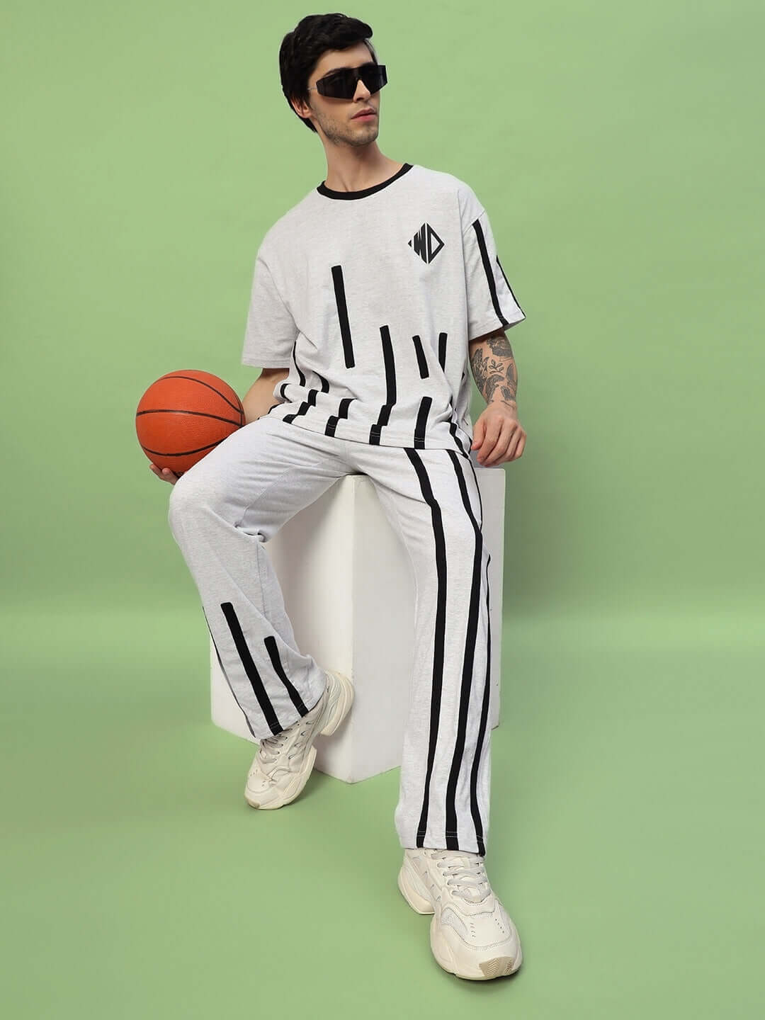Man in striped basketball-themed outfit sitting with a basketball on a green background, wearing sunglasses and white sneakers.