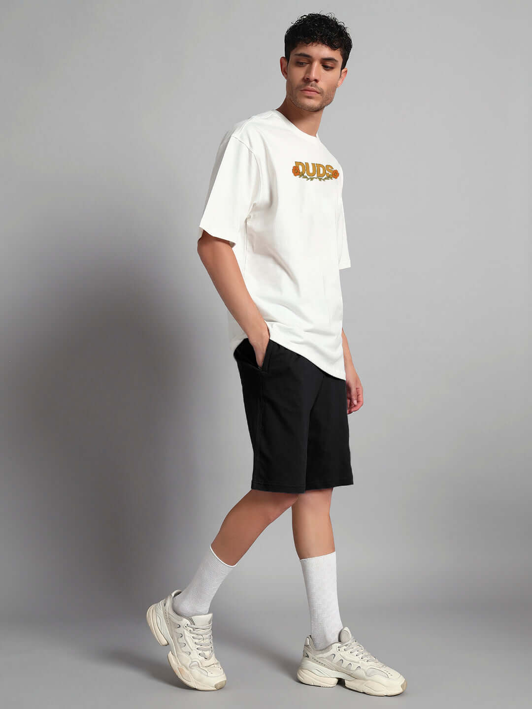 Man in casual outfit with white shirt and black shorts, posing against grey background, wearing sneakers and socks.