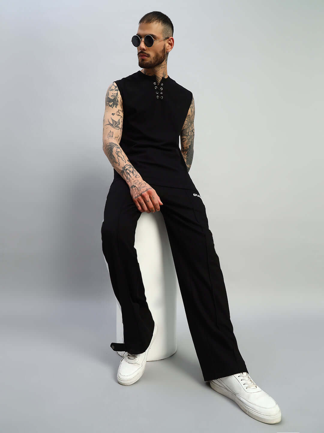 Person wearing black sleeveless top and black joggers with white sneakers, sitting on a white stool against a gray background.