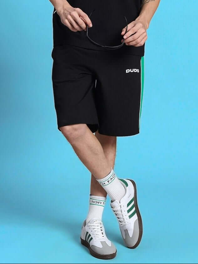 Person wearing METEORIC REGULAR FIT SHORTS BLACK showcasing casual style with socks and sneakers on blue backdrop.