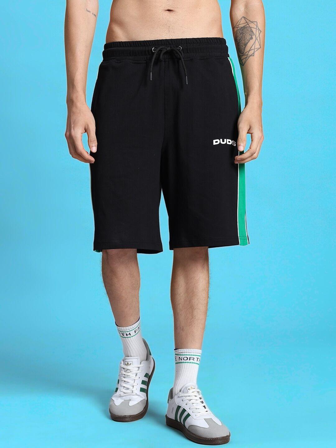 Person wearing METEORIC REGULAR FIT SHORTS BLACK with green stripe detail against blue background, showcasing casual style.