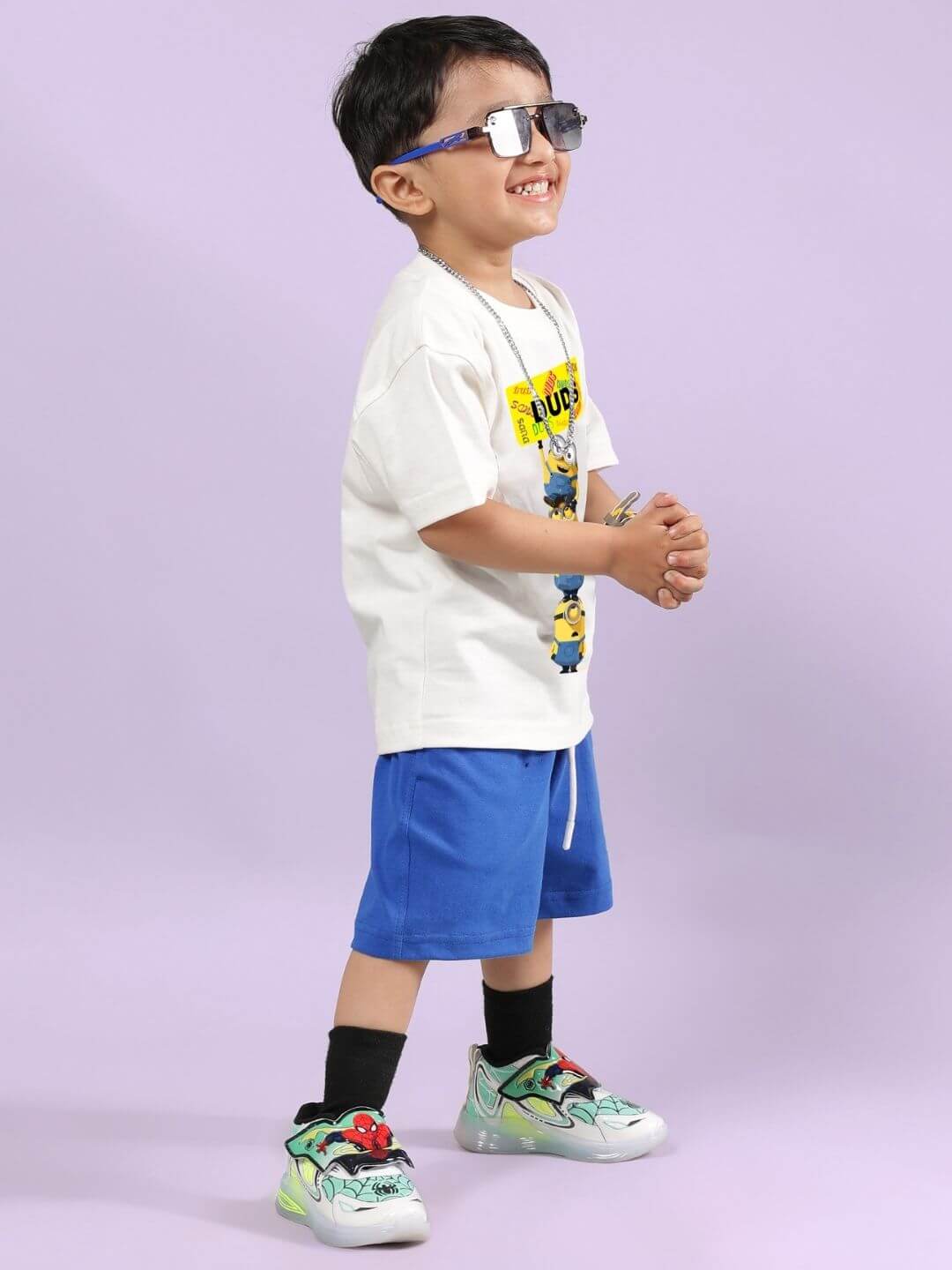 Boy wearing Minion cartoon T-shirt and blue shorts set, smiling and posing in sunglasses on a purple background.