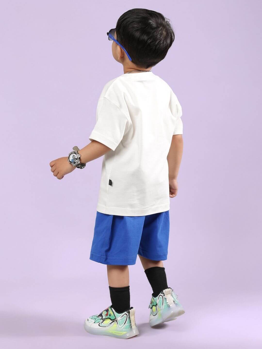 Back view of child in off-white T-shirt and blue shorts on pastel background, wearing colorful sneakers and sunglasses.