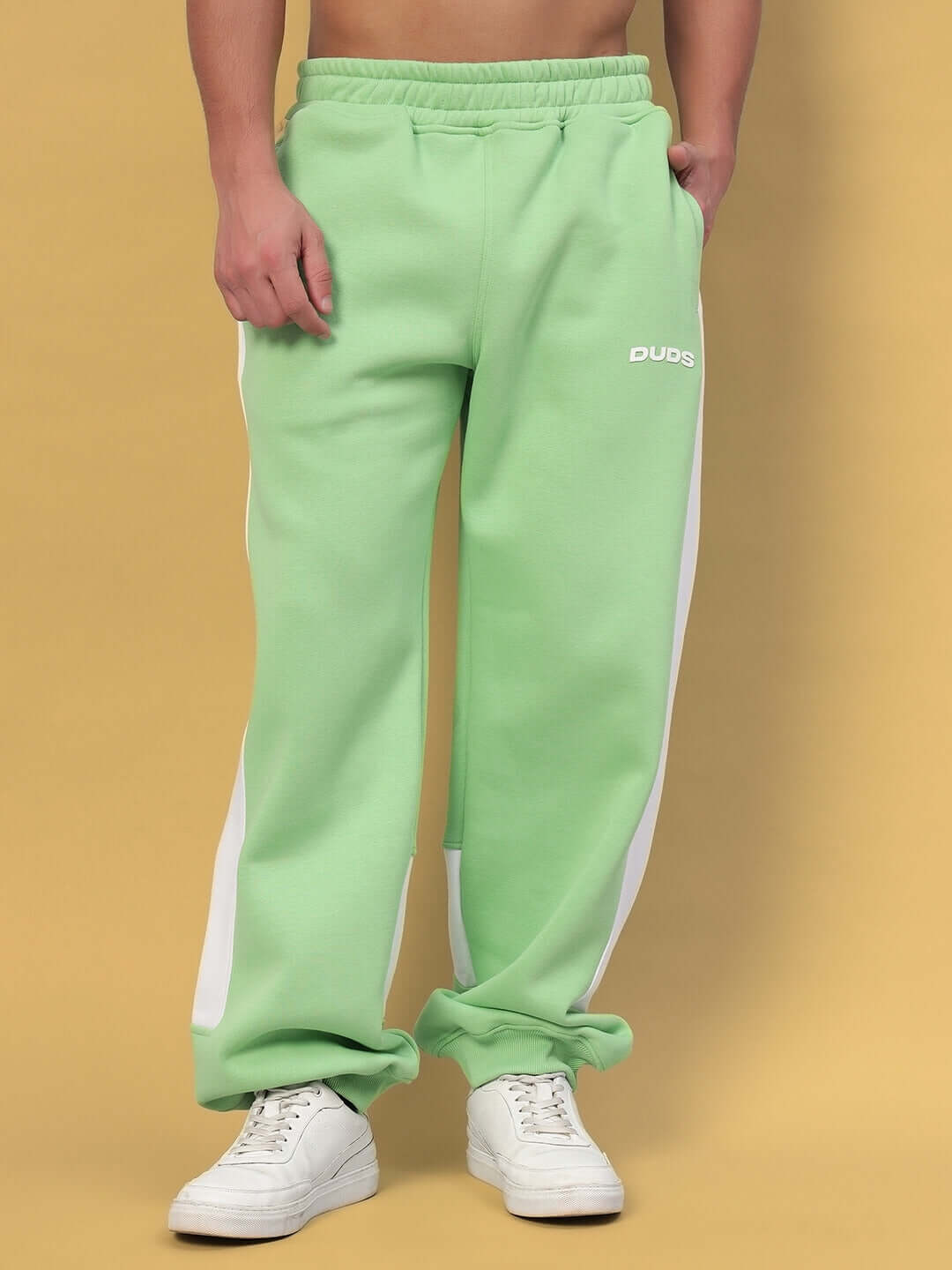 Person modeling MINTED BLISS FLEECE COLORBLOCK CO-ORD GREEN WHITE joggers with white sneakers on a yellow background.
