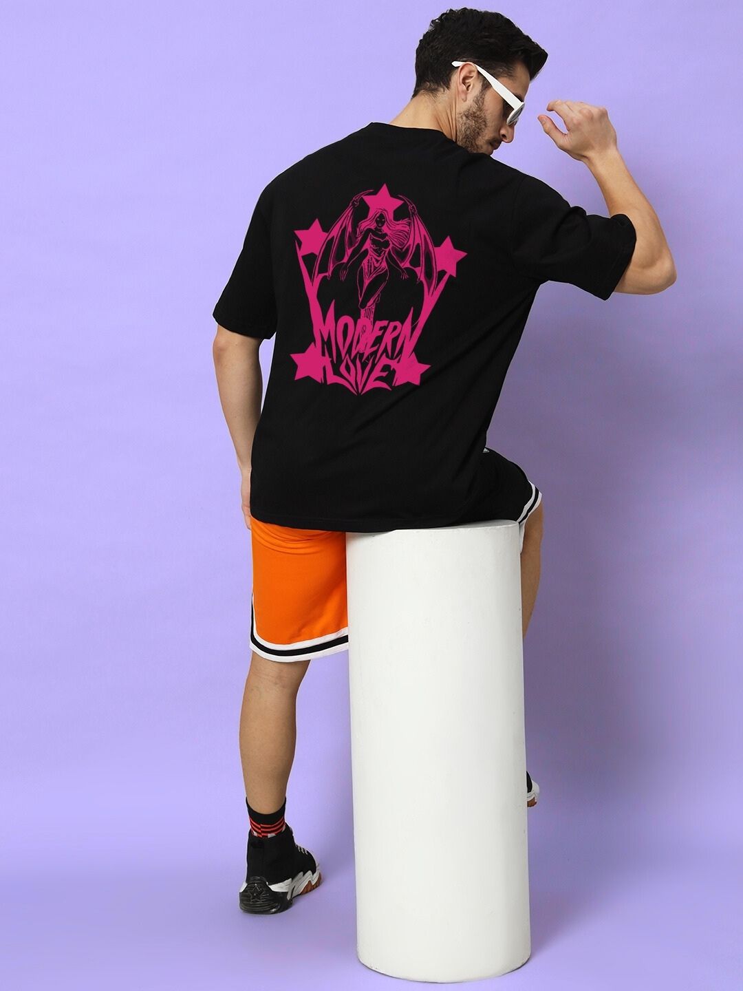 Rear view of model wearing MODERN LOVE OVER-SIZED T-SHIRT BLACK with pink graphic, seated on a white pedestal against a purple background.