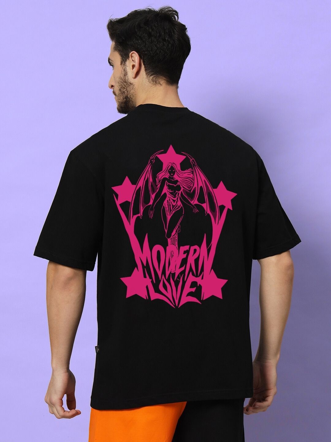 Model wearing MODERN LOVE OVER-SIZED T-SHIRT BLACK with pink graphic on back, styled against purple backdrop.