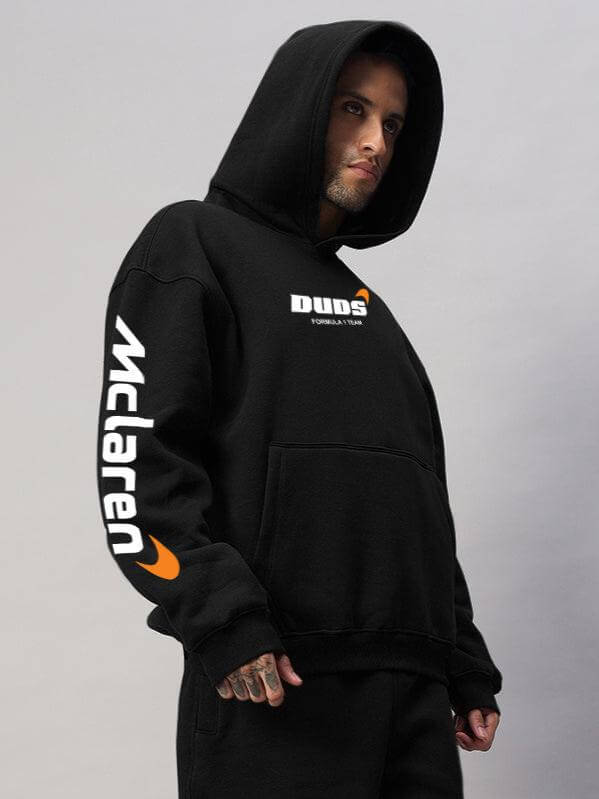 Male model wearing MONSTER DRINK FLEECE HOODIE BLACK with hood up, showcasing logos on arm and chest.