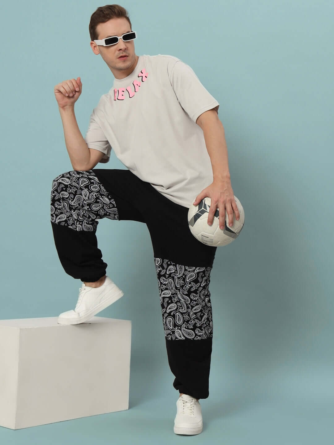 Model wearing black mosaic baggy joggers with white sneakers, holding a football against a blue background.