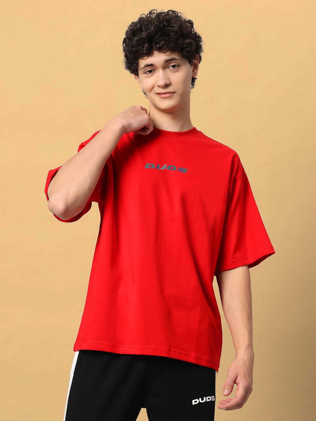 Young man wearing a red over-sized T-shirt featuring the MUDFLAP GIRL design by DUDS, styled casually against a neutral background.