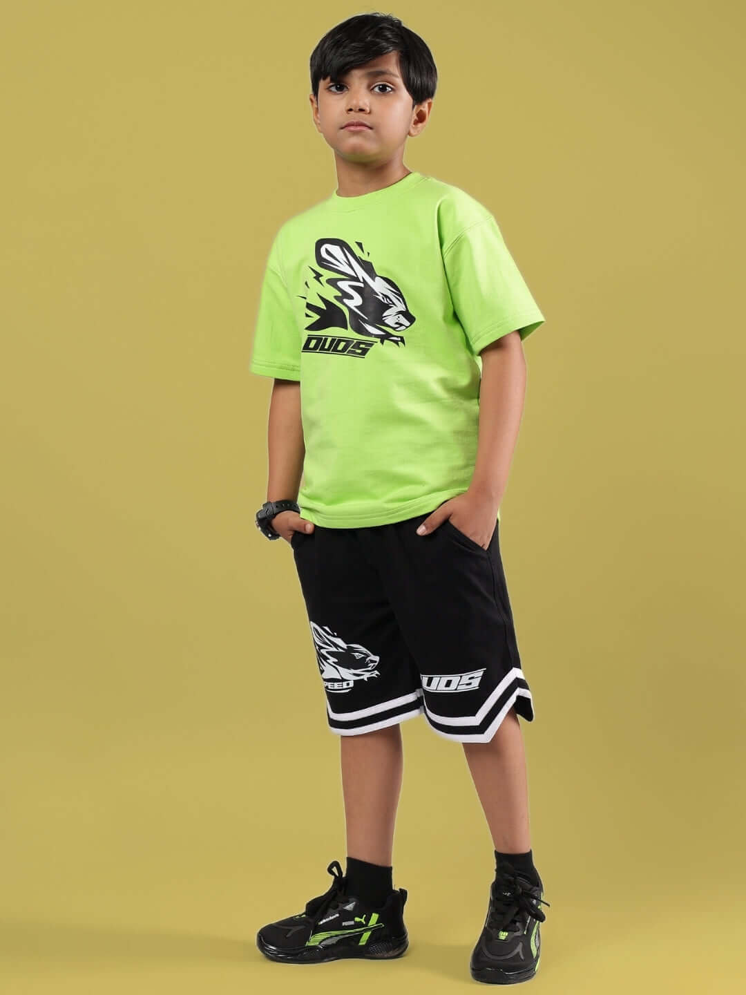 Child wearing MUFFIN SHORTS FOR BOYS & GIRLS BLACK with green Duds t-shirt, standing against yellow background.