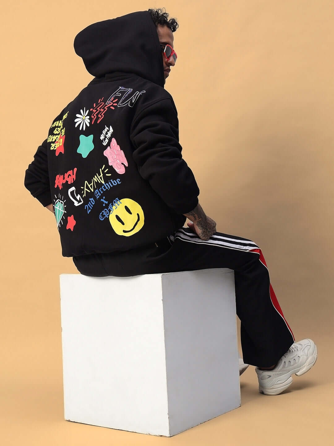 Person wearing NEVER BE AFRAID FLEECE HOODIE BLACK with colorful graphics, sitting on cube, showcasing street style fashion.