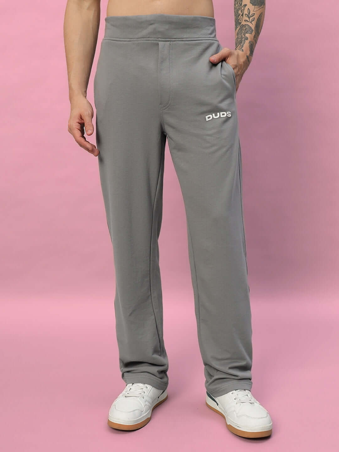 Man wearing grey DUDS relaxed fit joggers with white sneakers against a pink background.