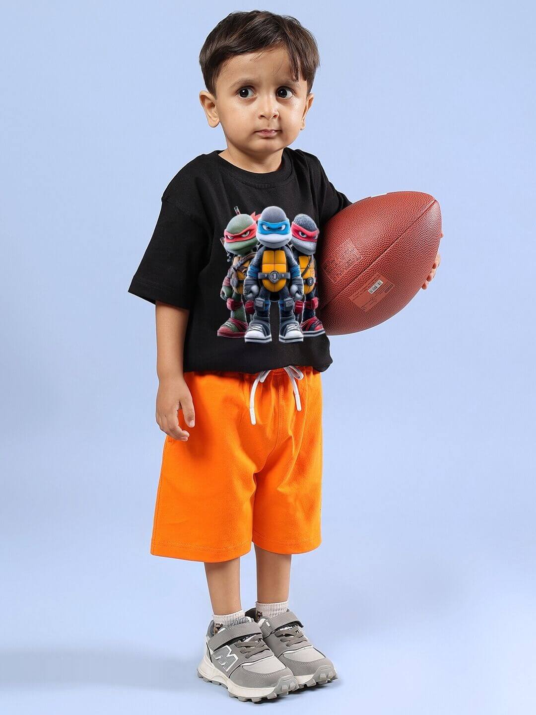 Young child wearing Ninja Turtles black T-shirt and orange shorts, holding a football, showcasing the fun co-ord for boys and girls.