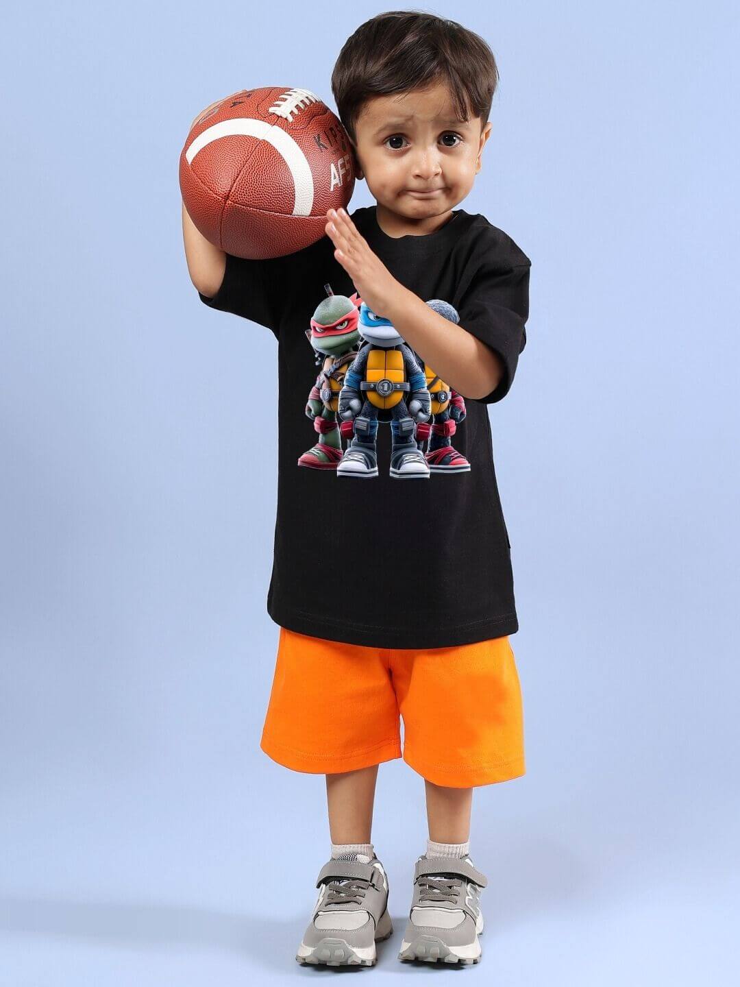 Child wearing Ninja Turtles co-ord set with black T-shirt and orange shorts, holding a football.
