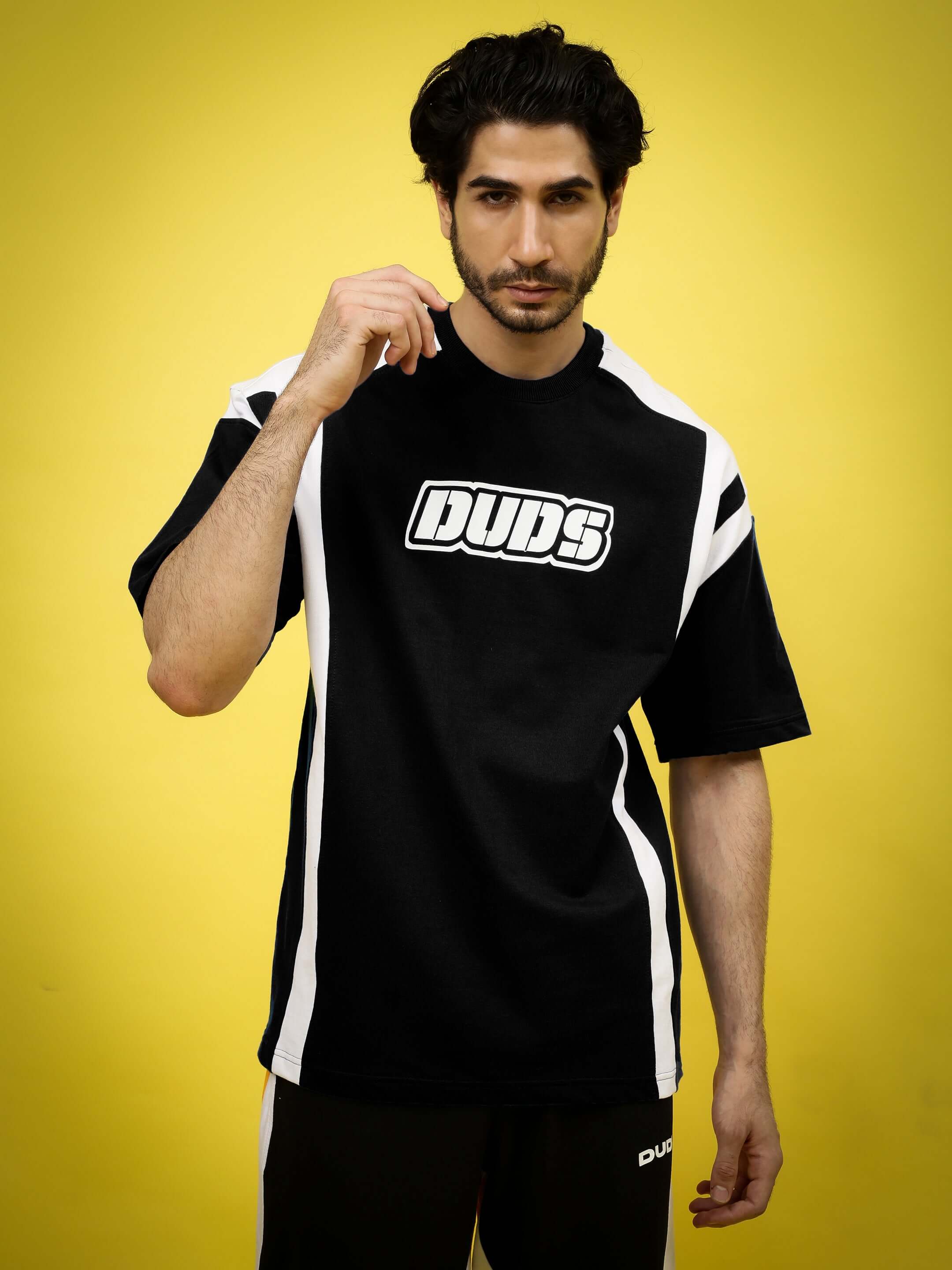 Model wearing a black and white oversized t-shirt with 'DUDS' logo, featuring durable cotton blend fabric and sporty design, against a yellow background.