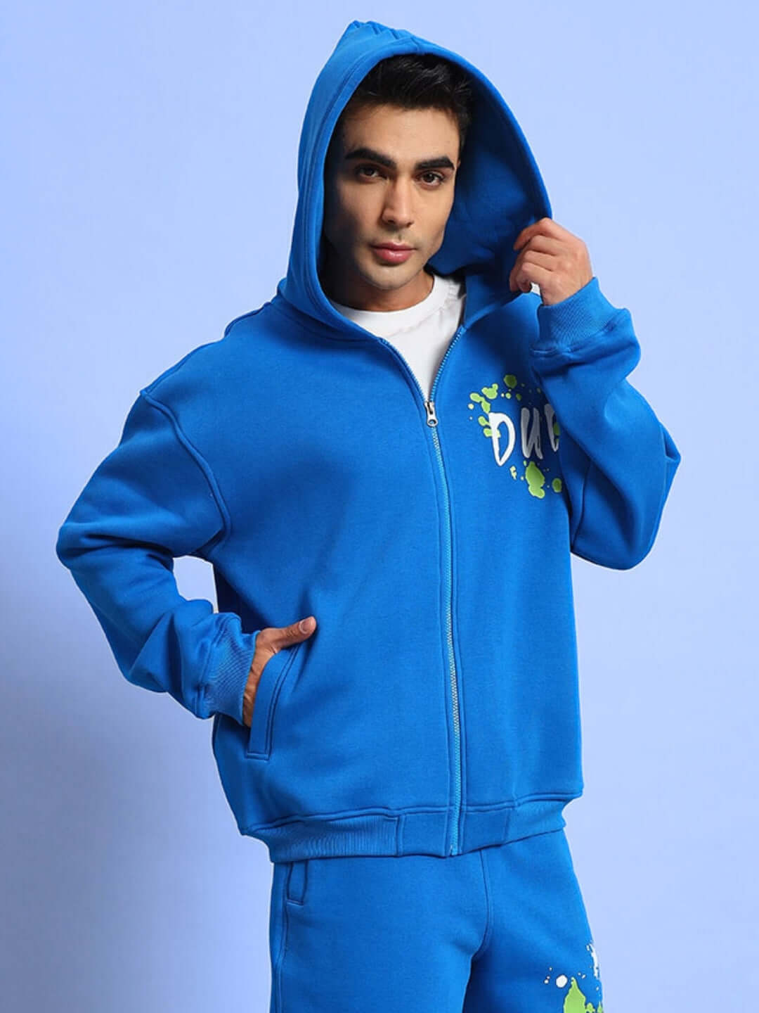 Model wearing blue No Drama cotton fleece hoodie with hood up, featuring artistic splashes and zipper detail.