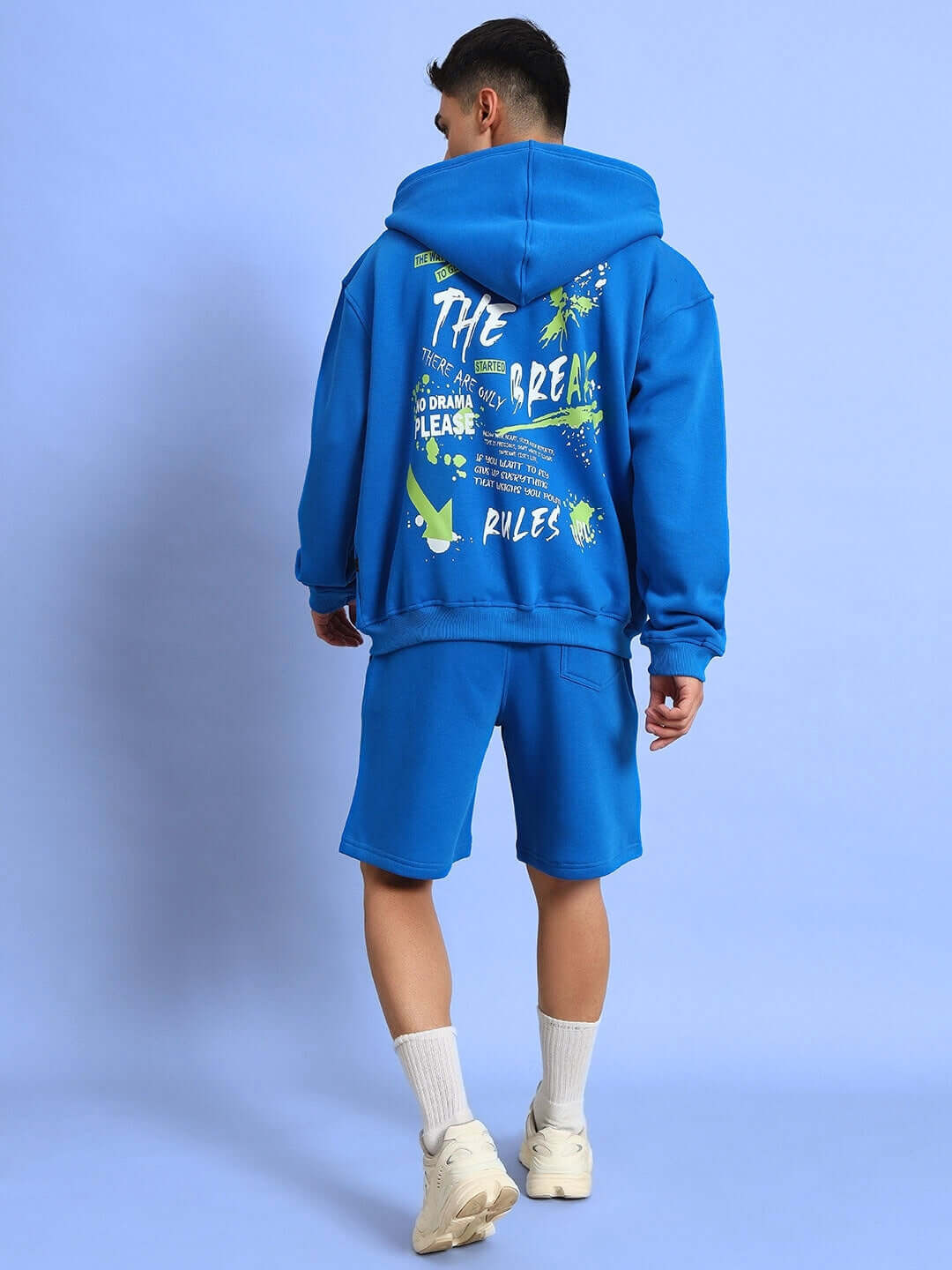 Man wearing blue No Drama Fleece Co-Ord set with graphic back design, featuring hoodie and shorts, on a light blue background.